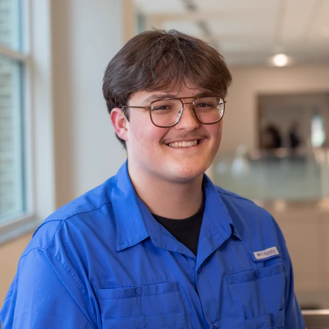 A young man with glasses and a mustache smiles while wearing a royal blue Columbia shirt. He is standing indoors next to a window, with soft natural light illuminating his face and a blurred hallway in the background.
