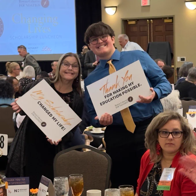 Students pose with thank-you signs at a scholarship luncheon event. One student in the center wears a blue shirt and yellow tie and holds a sign that says “Thank You for Making My Education Possible.” The room is full of seated guests enjoying lunch, with a large presentation screen in the background displaying the event title “Changing Lives Scholarship Luncheon.”