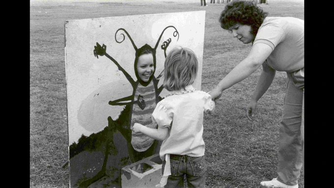 Black and white photo of a young child outside looking at a woman whose face is in a cutout painting of a bug. 