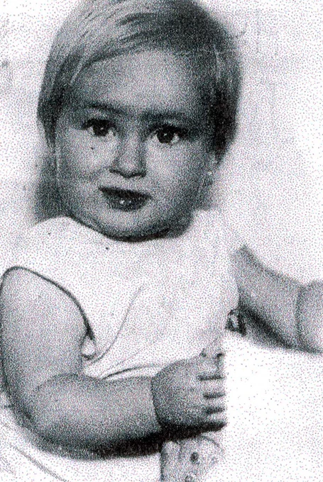 A vintage black-and-white photo of a young girl with a focused expression, wearing a light sleeveless top. The child's hair is neatly combed, and one hand is slightly raised.
