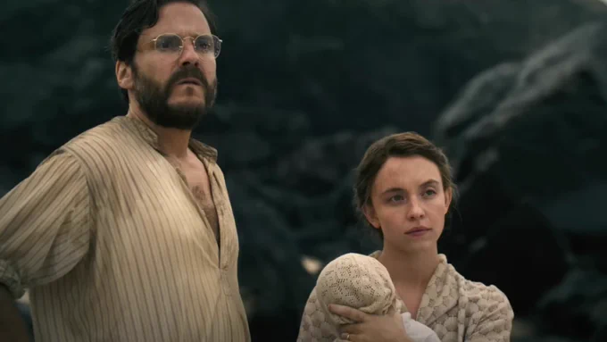 Daniel Brühl (left) and Sydney Sweeney (right) standing with a baby in front of rocks on a beach looking at something approaching off in the distance in a still from writer-director Ron Howard's new film EDEN. 