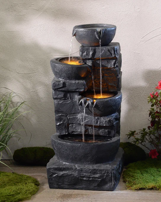 A decorative tiered water fountain illuminated with warm lighting, surrounded by lush greenery and a vibrant red flower, set against a textured grey wall.