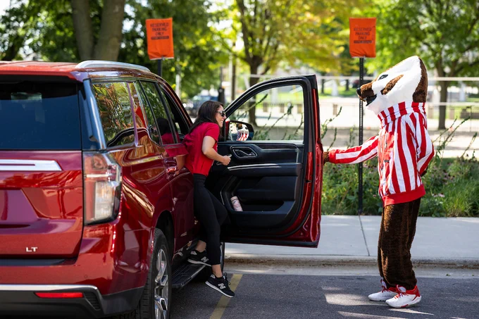 Mascot Bucky Badger opens a vehicle door for a parent while welcoming new students moving into University Housing.