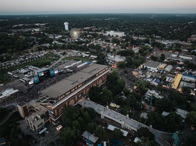 Aerial view of the Minnesota State Fairgrounds crowded with visitors, featuring prominent buildings and a lit Ferris wheel in the background.