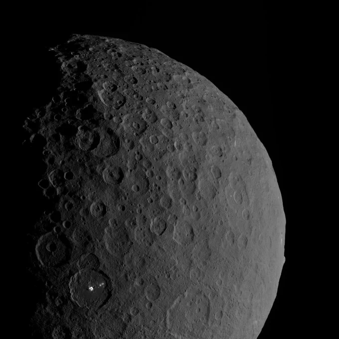 The gray, cratered surface of Ceres fills most of the frame. Near the bottom left, one crater has small patches of much brighter material.