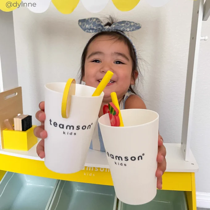 A child smiles while holding two cups with straws, branded with "teamson kids," in front of a lemonade stand.