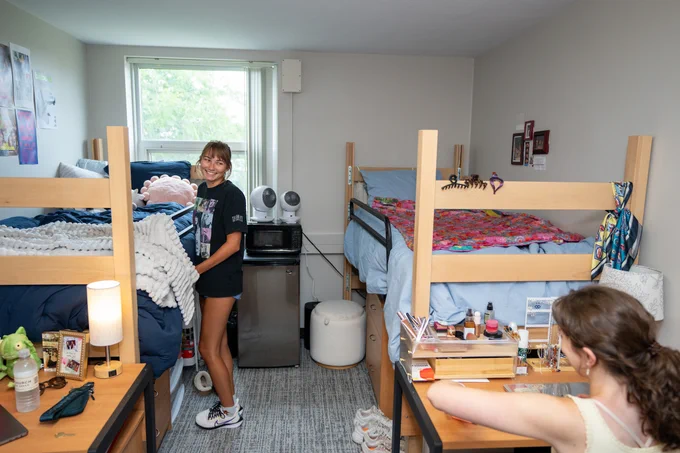 Two people in a college dorm room with two bunk beds, desk areas, and a window. The room is filled with personal items and decorations, making it cozy and lived-in.