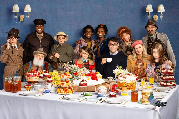 A photo showing 13 performers in casual, vintage, outfits sat at a dining table laden with pastry treats 