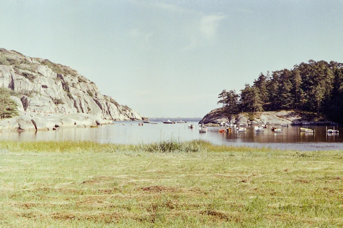 A small fjord between a mountain and a forest, seen from a grassy shore. Boats and people swimming in the water, sunbathing people on the shore. Bright, sunny weather and blue sky.