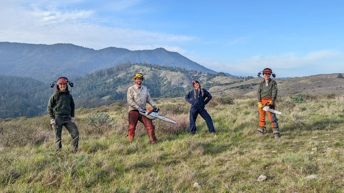 four people wearing safety equipment holding tools pose in front of a mountain.