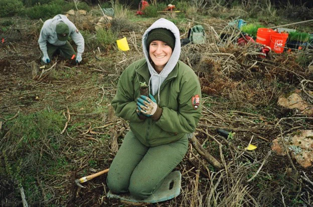 A smiling person stops pulling weeds to pose for a photo. There is another person and buckets behind them.