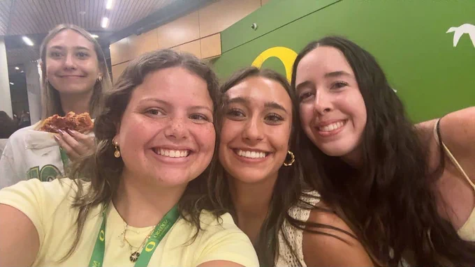 Four people smiling at the camera in a room with a green logo in the background. One person is holding a slice of pizza.