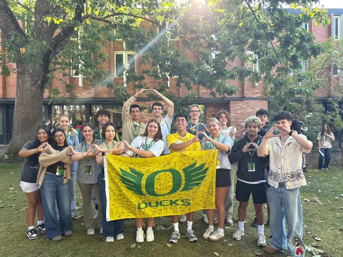 Group of individuals throwing the O with their hands, holding a University of Oregon Ducks flag outdoors on a sunny day.