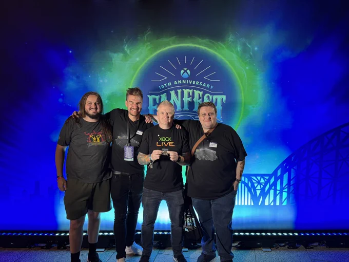 Craig Duncan with Cats, Leator and Loki in front of a colorful Xbox FanFest 10th Anniversary backdrop featuring a cosmic design and the Cologne skyline. Craig is displaying a special FanFest edition Obsidian Underpants coin, created and gifted by us.