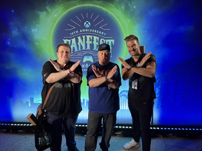 Aaron Greenberg with Leator and Loki posing with arms crossed in an ‘X’ in front of a colorful Xbox FanFest 10th Anniversary backdrop featuring a cosmic design and the Cologne skyline.