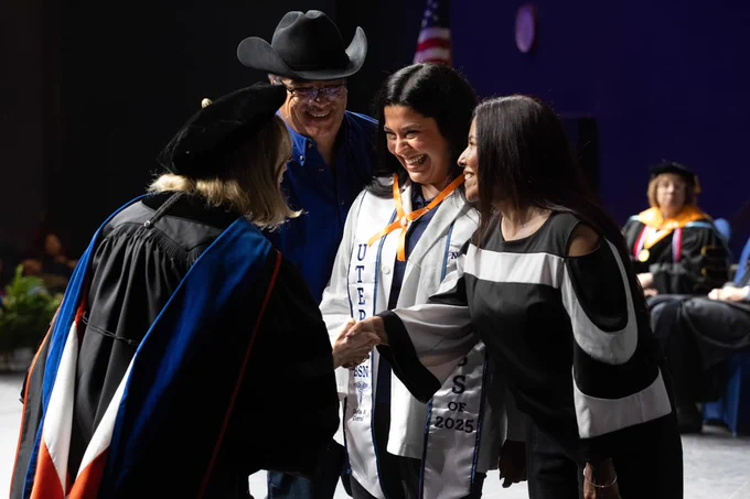Graduate in white sash marked 'UTEP Class of 2025' is congratulated by university officials at a pinning ceremony, with attendees smiling and a U.S. flag in the background.
