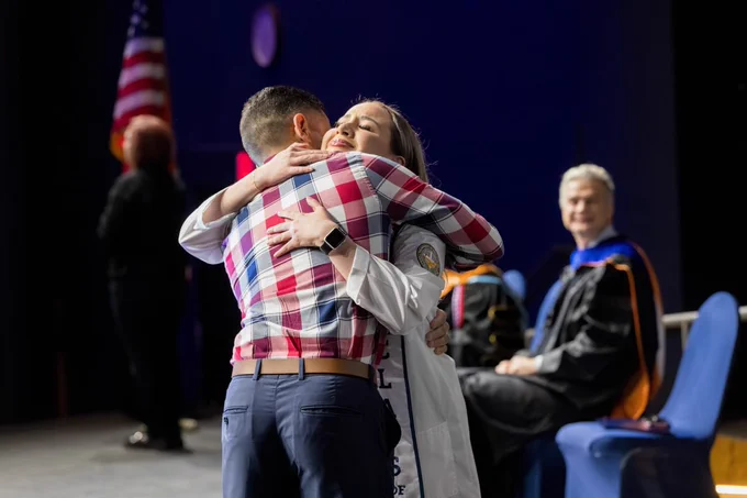 Two individuals embrace warmly on a stage at a pinning ceremony, with one donning a white coat. An American flag and a seated academic figure appear in the background.
