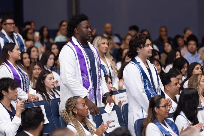 Students wearing white coats and honor cords at a nursing pinning ceremony, seated and standing in an auditorium.
