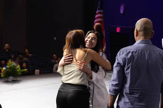 Two individuals embracing joyfully on stage at the UTEP Nursing Pinning Ceremony, with an audience and the American flag in the background. One individual is wearing a white coat. 
