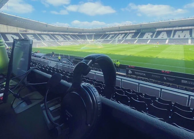 A view from the media press bench at Pride Park Stadium, home of Derby County Football Club. This image shows a pair of headphones resting on a desk with a computer monitor nearby, overlooking the football pitch. The stadium is empty, with the sun shining brightly, and the words ''THE RAMS'' are visible on the seats opposite. The grass is well-maintained.