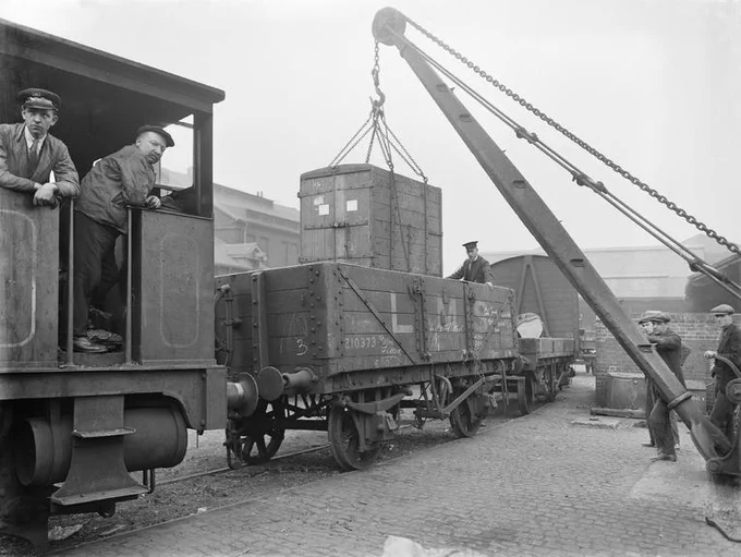 Crane being used to load a railway wagon with a container box; railwayman standing in the wagon to guide the load down. A shunting engine is coupled to the wagon near the camera, with the footplate crew looking on.
Image courtesy National Railway Museum