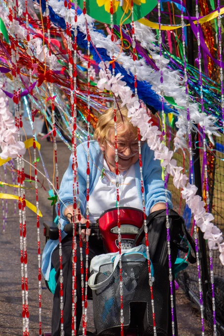 A brilliantly colourful image from our super sensory that shows a lady on a red mobility scooter wearing a white t shirt with a light blye cardigan over the top. She has what looks like a nasal cannula and has the largest grin on her face as she drives through hanging beads that are red and white and glittery streamers in all sorts of colours. 