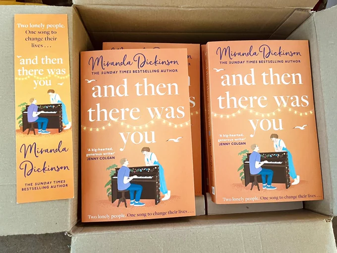 A box containing paperback editions of my new novel, And Then There Was You. The cover shows a man sitting at a painted piano with a woman leaning against it, listening to him play. Strings of white lights loop above with two seagulls flying. The tagline reads: “Two lonely people. One song to change their lives.”