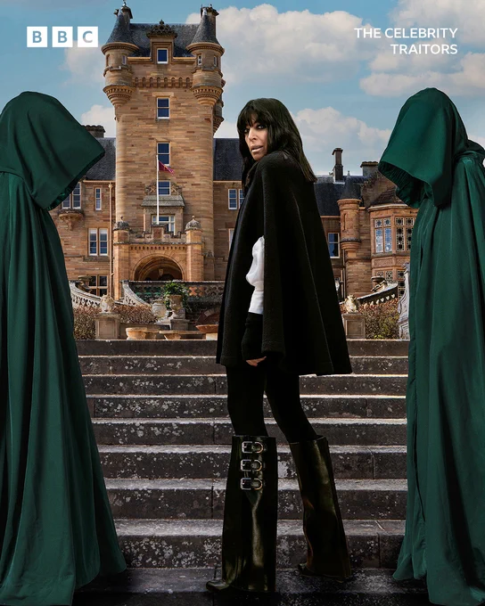 Claudia Winkleman stands on the steps outside the Traitors castle dressed in black, flanked by two figures in green hooded cloaks.