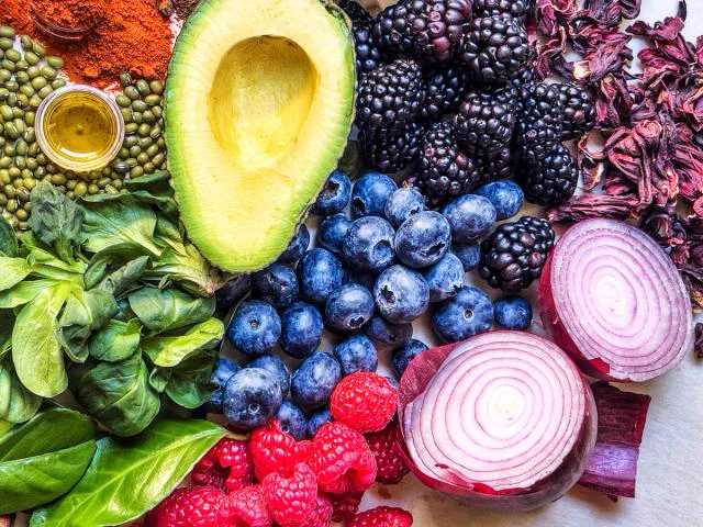 Various fresh foods including avocado, blackberries, blueberries, raspberries, red onion slices, green lentils, olive oil, and basil leaves arranged on a flat surface.