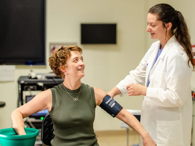 Lacy Alexander sits in a chair with her hand in a bucket of ice water, simulating a patient in the experiment. Auni Williams stands beside her, monitoring Lacy's blood pressure. 