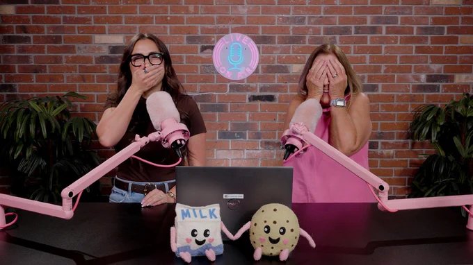 Two individuals standing at a desk with pink microphones, covering their mouths with their hands in a playful pose. They are in front of a brick wall with a neon sign reading "LIVE ON AIR." A laptop and two plush toys labeled "MILK" and "COOKIE" are on the desk.