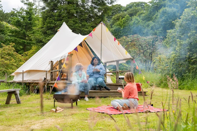 Three women sit outside a bell tent in the grounds of a castle. Visit Lake District/Paul Mitchell