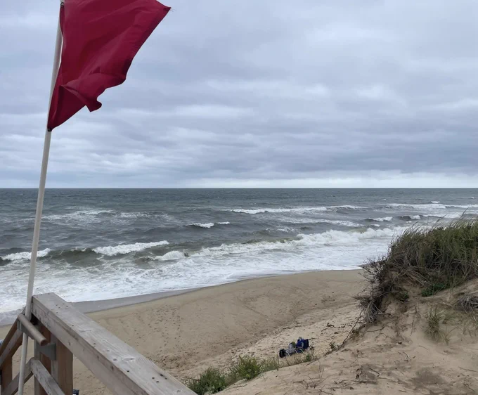 Large ocean waves lap up on the sandy beach. Red flag whips in the wind.