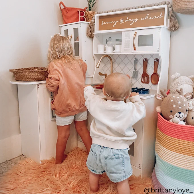 Two children playing with a toy kitchen set, with text "sunny days ahead" displayed above the setup.