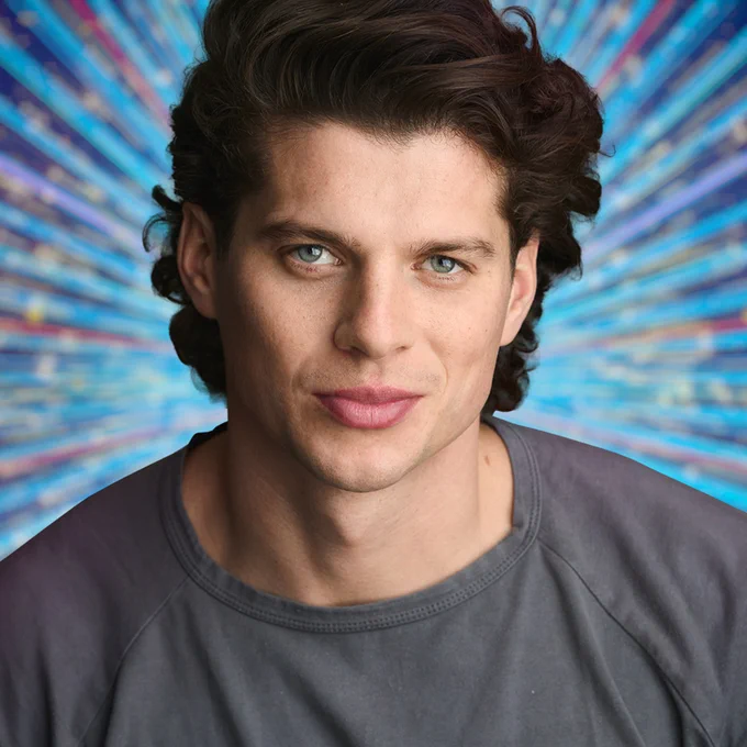 A young man in a grey t-shirt smiles for the camera, set against a glittering blue background
