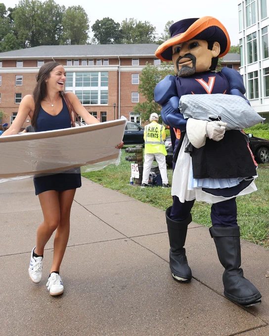 A person in casual clothing carrying a large silver tray laughs while walking alongside the University of Virginia's mascot, Cavman, who is dressed in a blue and orange outfit.