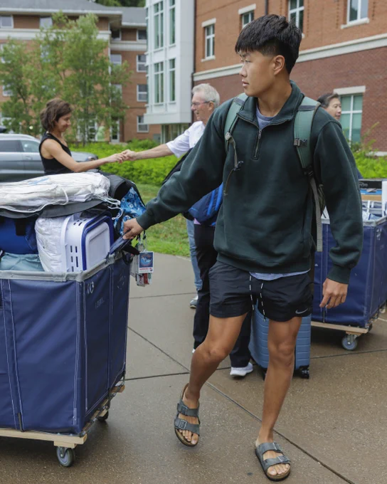 A student pushes a heavily loaded cart across a campus during move-in day, with other people and residential buildings in the background.