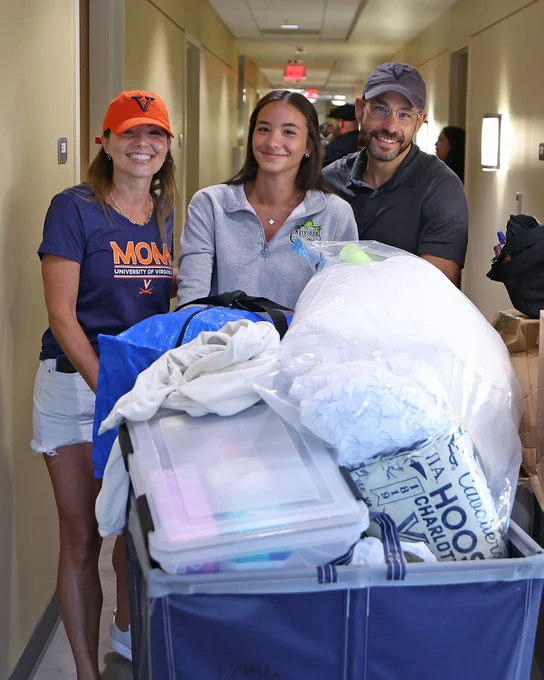 Three people smiling in a hallway, surrounded by moving items in a blue cart, with one person wearing a "Virginia" cap and another wearing a "Mom UVA" shirt.