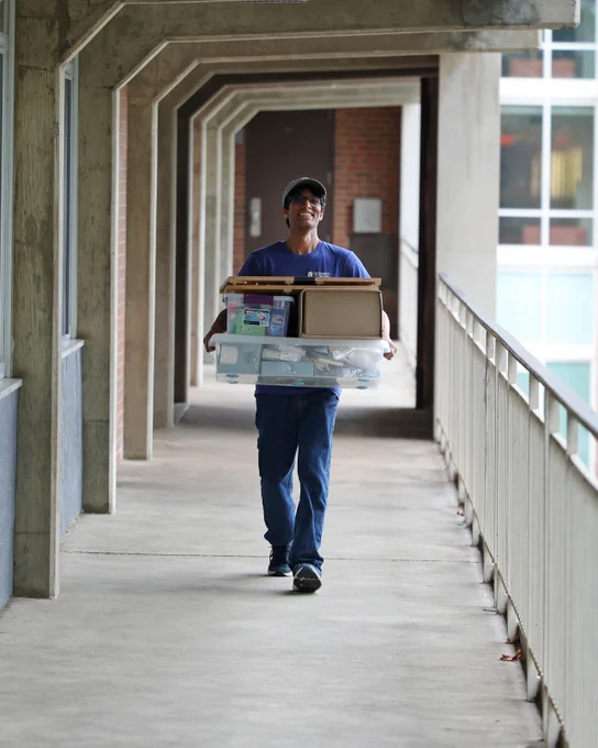 A person in a blue shirt and blue pants carries multiple boxes while walking along a covered walkway, smiling.