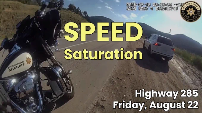 A Jefferson County Sheriff’s Office motorcycle is parked on the side of a highway next to a white SUV that has been pulled over. The road is bordered by grassy hills and mountains under a partly cloudy sky. Overlay text reads “SPEED Saturation” in large yellow letters. Additional white text at the bottom right corner says “Highway 285, Friday, August 22.” The Jefferson County Sheriff’s Office logo is visible in the top right corner.