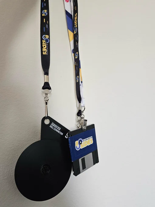 Two badges are displayed hanging from lanyards. One badge is a CD on a 3D-printed holder, and the other a floppy disk with a sticker displaying the BSides Bloomington logo wrapped around it. 