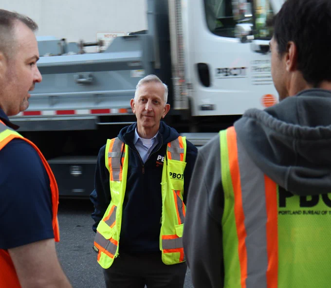 Mayor Wilson standing, facing two Portland Bureau of Transportation employees while wearing a yellow safety vest. There is a PBOT maintenance truck in the background.