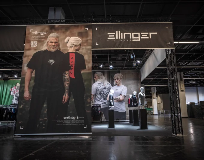 The entrance to a pop-up shop with Witcher merchandise. On the right is a floor-to-ceiling photograph of cosplayers with the hair and makeup of Geralt and Ciri, dressed in the shop’s contemporary merchandise. Above the entrance is a large Ellinger Clothing logo. Mannequins can be seen inside.