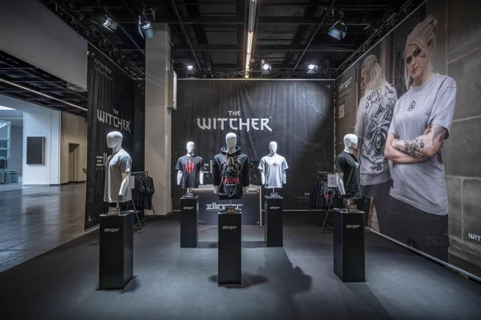 An empty pop-up shop with Witcher merchandise contains five mannequins, all wearing different Witcher-themed shirts or hoodies. The base of each mannequin shows the logo of Ellinger Clothing. On the right is a large-scale photograph of a man and a woman wearing the same merchandise, scaled up in size to serve as a wall.