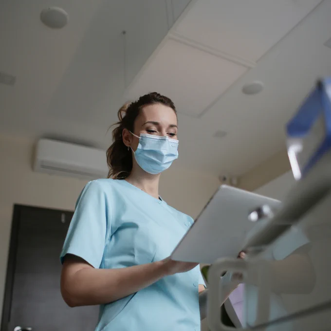 Healthcare professional in light blue scrubs holding a tablet in a clinical setting with modern equipment.