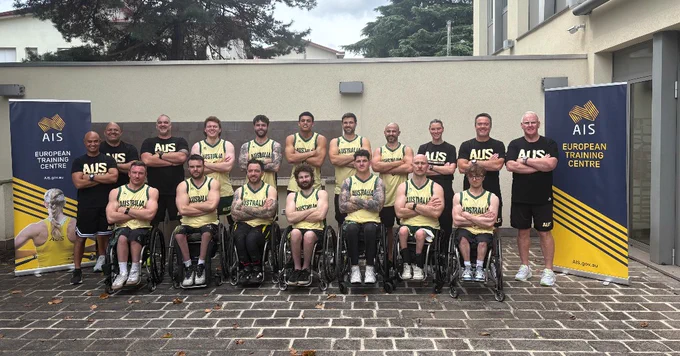 Group photo of the Australian wheelchair basketball team at the AIS European Training Centre, with athletes in team uniforms seated in wheelchairs, smiling.