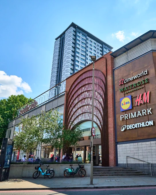 Exterior view of the Southside Shopping Centre in Wandsworth, London featuring a curved red facade with signs for Cineworld, H&M, Primark, and Decathlon. A tall residential tower stands in the background under a clear blue sky.