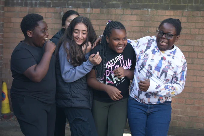 Group of five teenagers standing close together outdoors against a brick wall and laughing. 