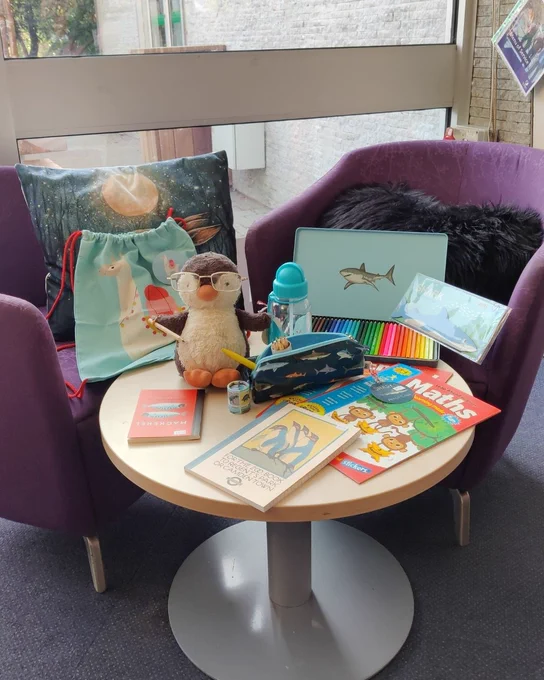 Photograph of a circular coffee table in front of two purple chairs. Displayed on the table is a penguin toy surrounded by notebooks, workbooks, coloured pencils, a pencil case, a water bottle, & a drawstring bag. 