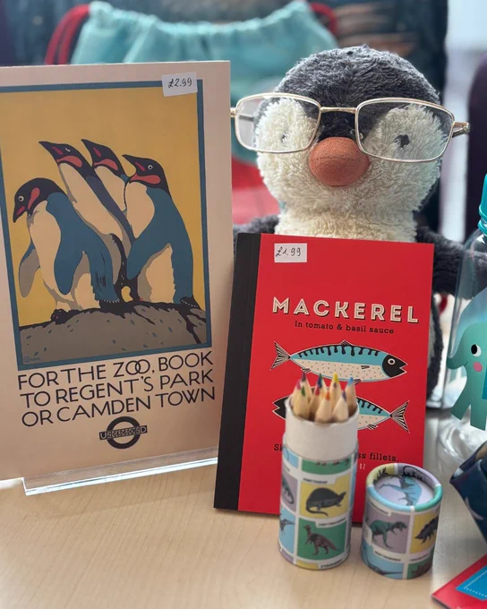Photograph of a penguin toy holding a small red notebook with mackerel on it. In front of the notebook is a pot of coloured pencils with a dinosaur pattern & the lid off next to it. Next to the penguin is a notebook with penguins on it. 